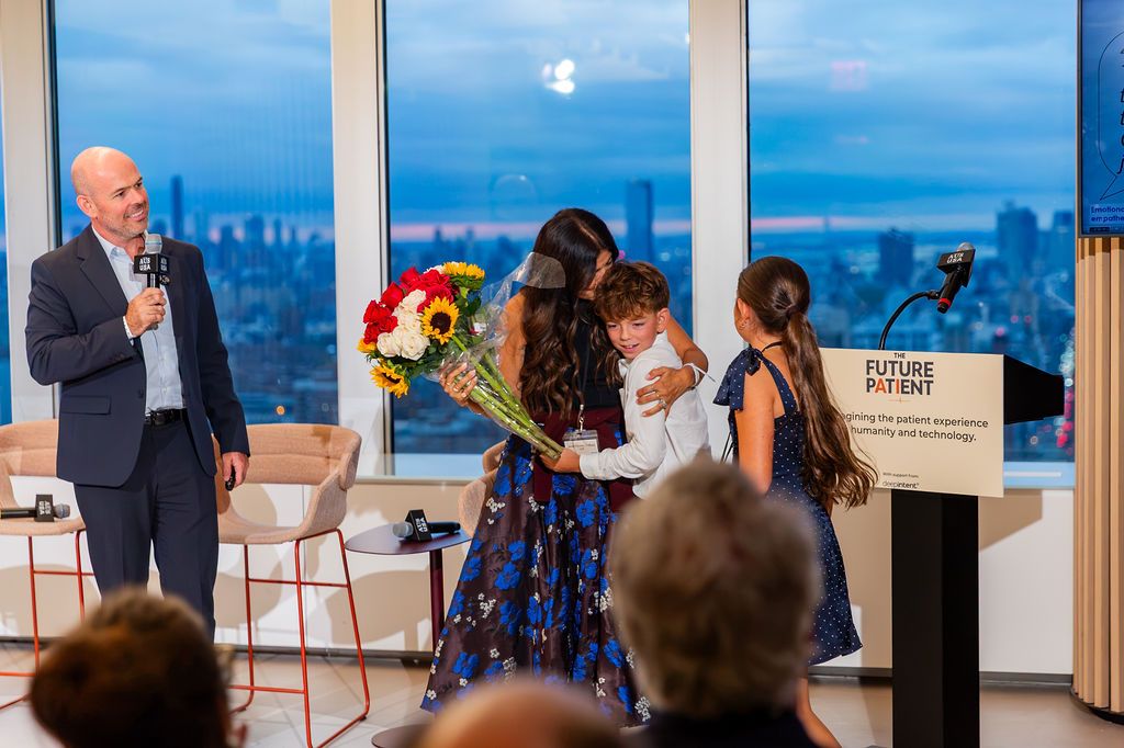 Janet Gomez holding a bouquet and hugging their son, their daughter beside her, John speaking at the microphone.