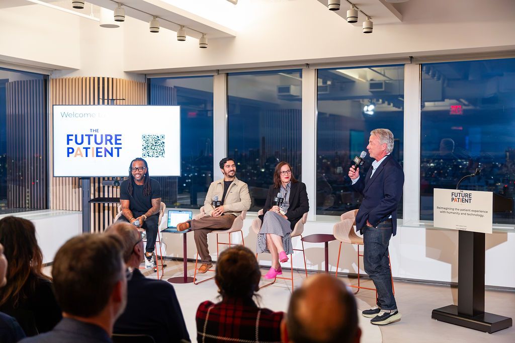 The panel on stage in front of a full audience, the New York skyline behind them and a screen reading Welcome to The Future Patient.