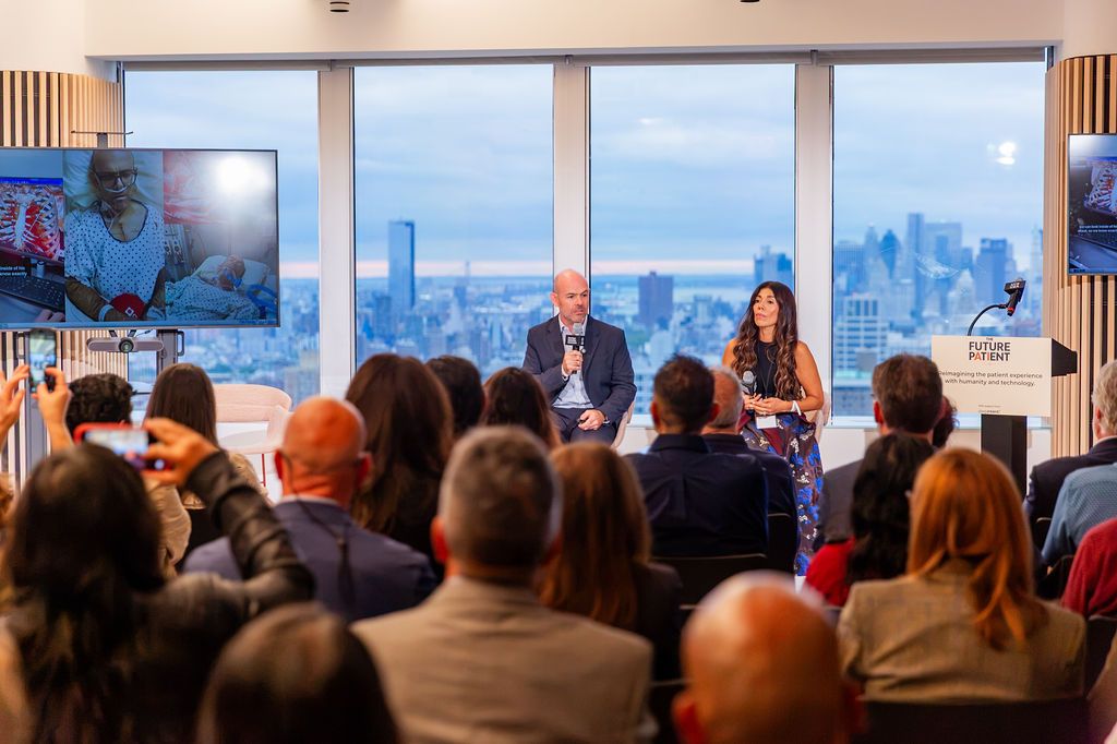 John Duffield and Janet Gomez on stage with footage from the ICU on the screen beside them, the audience watching from a packed room.