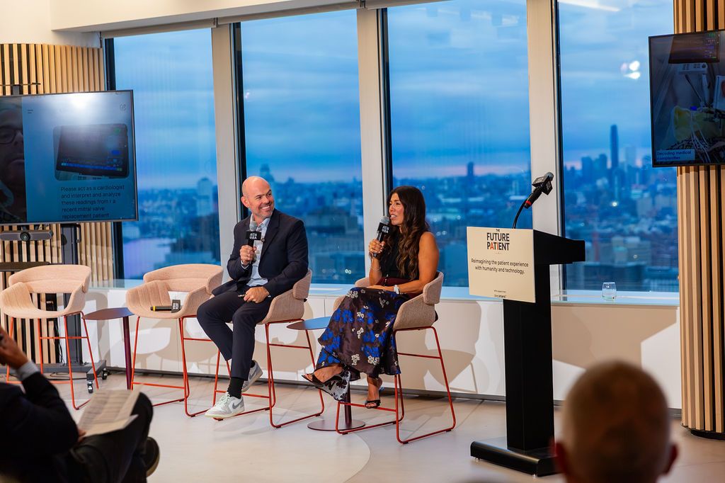John and Janet seated on stage, a screen behind them showing an AI prompt asking a model to interpret readings from a mitral valve replacement patient.
