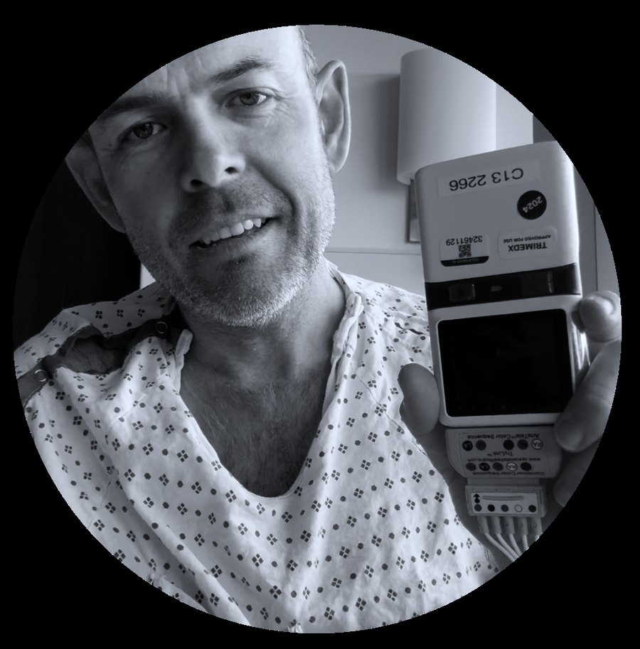 Black-and-white portrait of John Duffield in a hospital gown, holding a cardiac telemetry monitor.